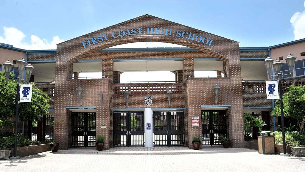 A modern school building with brick walls and glass doors. Above the entrance, a sign reads "First Coast High School".