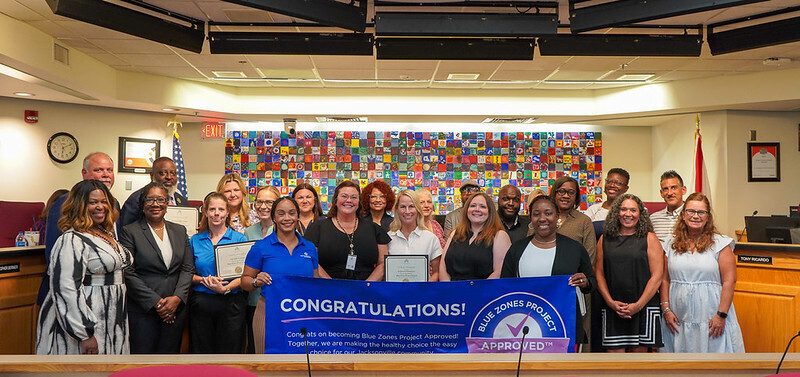 Group of people posing for a photo in a council chamber with a colorful mural behind them.