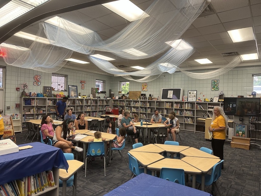 A group of parents and children in the media center with the principal standing in the front talking.