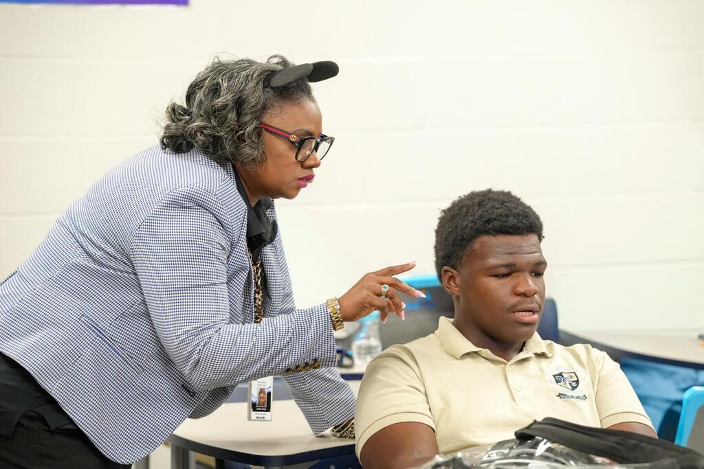 Woman in glasses and blazer pointing at a seated man in a yellow shirt, in a classroom.
