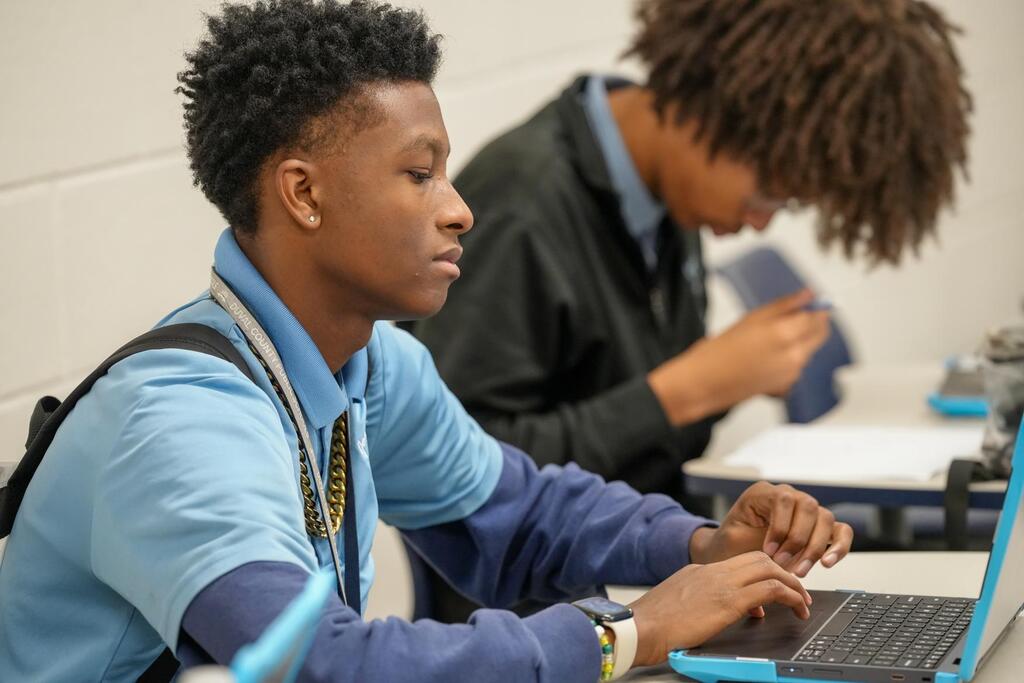 Two students in a classroom, one typing on a laptop and the other looking down.