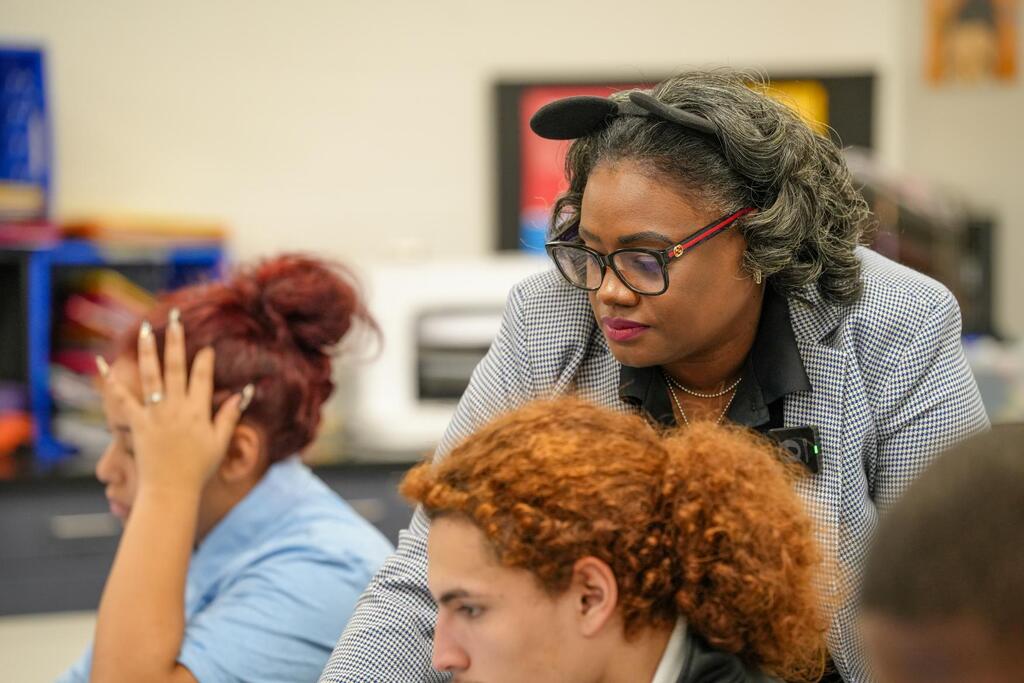 A teacher with glasses leans over a student with red curly hair. Another student with red hair touches her face.