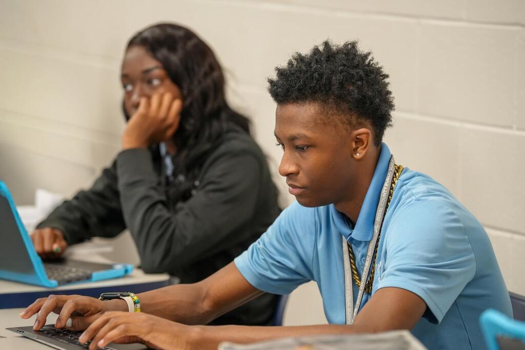 Two students seated at desks in a room with a white wall, one using a laptop, the other with hand on face.
