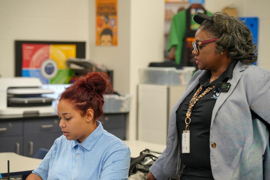 Two women in a classroom. One in blue is seated, and the other in gray is standing.