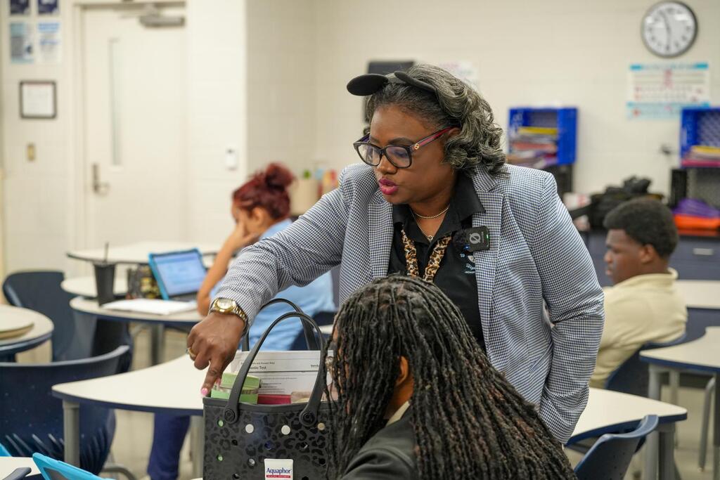 Woman standing near a student, holding a bag. Student with braided hair. Background includes desks, tables, chairs, and a clock.