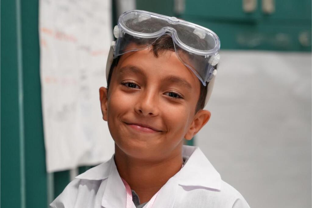 A smiling boy in a white lab coat and goggles. Behind him is a green wall and a piece of paper.
