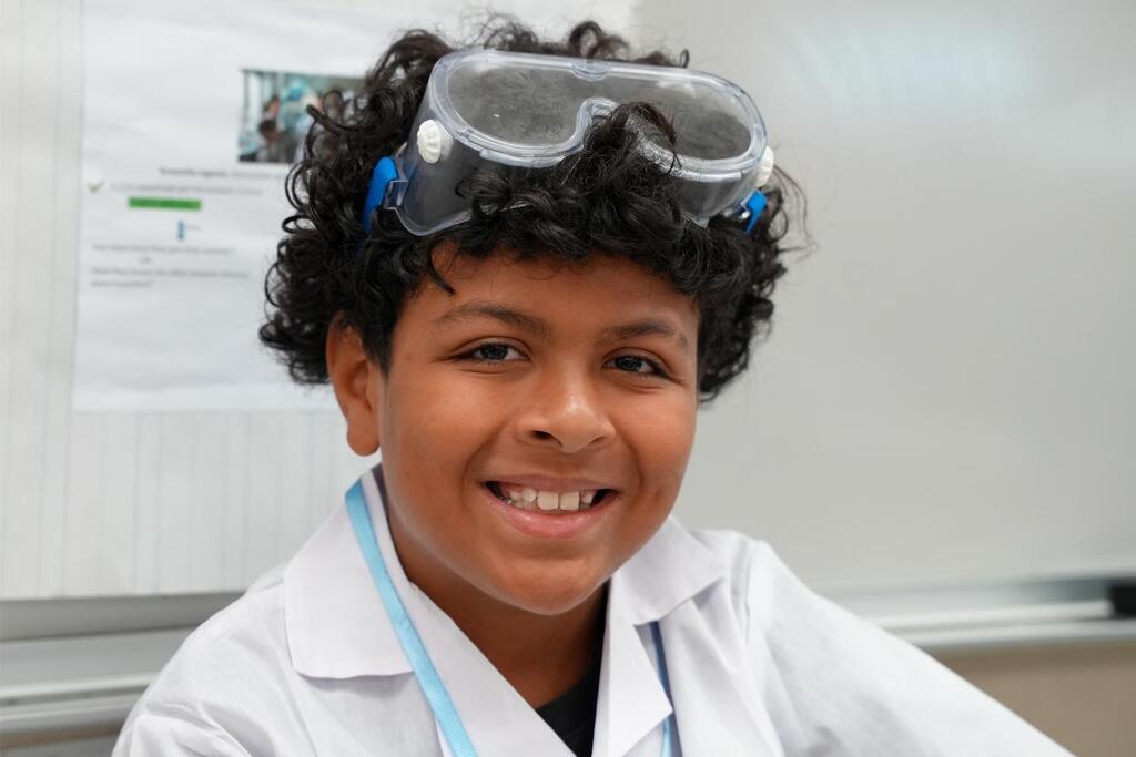 A smiling child in a white lab coat and safety goggles sits against a white board.
