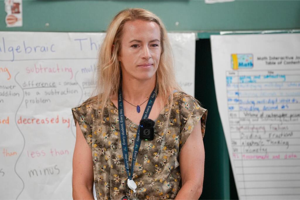 A woman with blonde hair stands in front of whiteboards with writing and drawings. She wears a floral blouse and a blue lanyard.