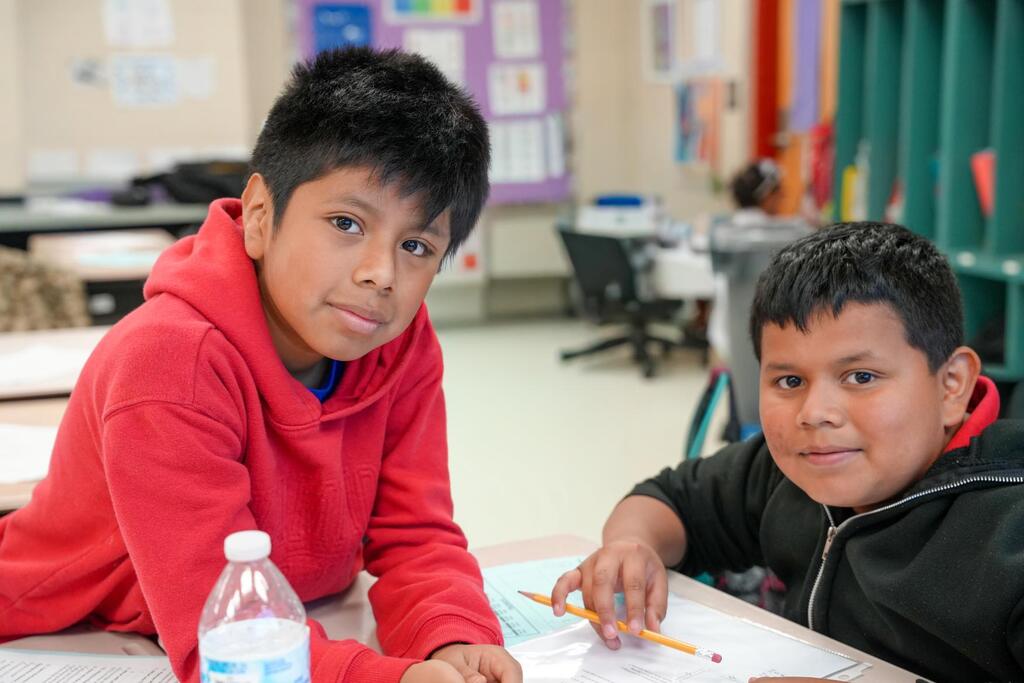 Two children seated at a table, one wearing a red hoodie and the other in a black jacket.