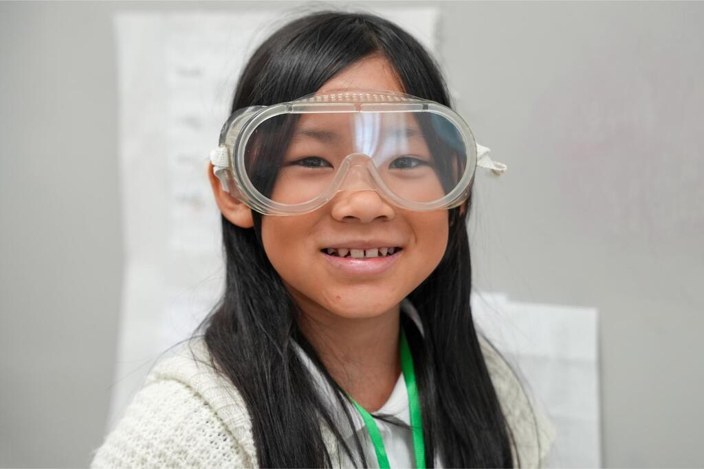 A girl with long dark hair wearing safety goggles, a white sweater, and a green lanyard against a light gray background.