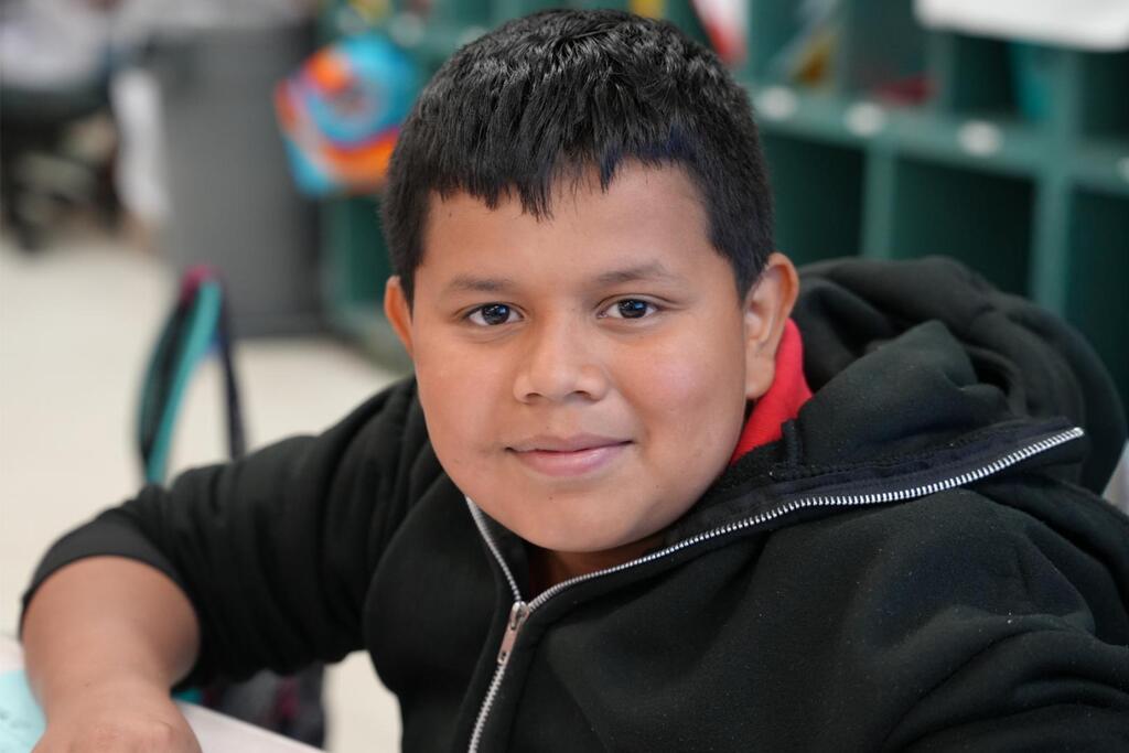A child with dark hair wearing a black hoodie with a red collar sits in a classroom.