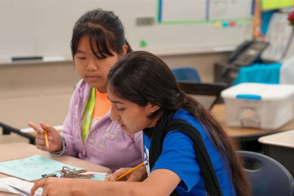 Two students at a desk, one holding a pencil and looking down, the other holding a pen and looking up.