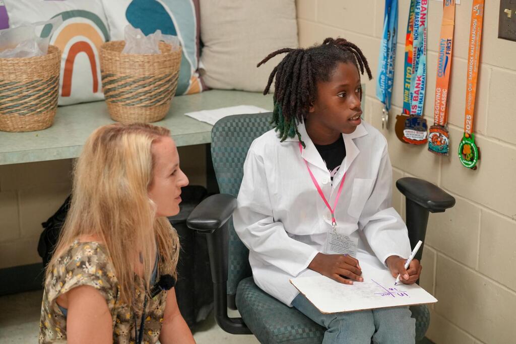 A person with long hair sits in a chair and talks to another person with blond hair.