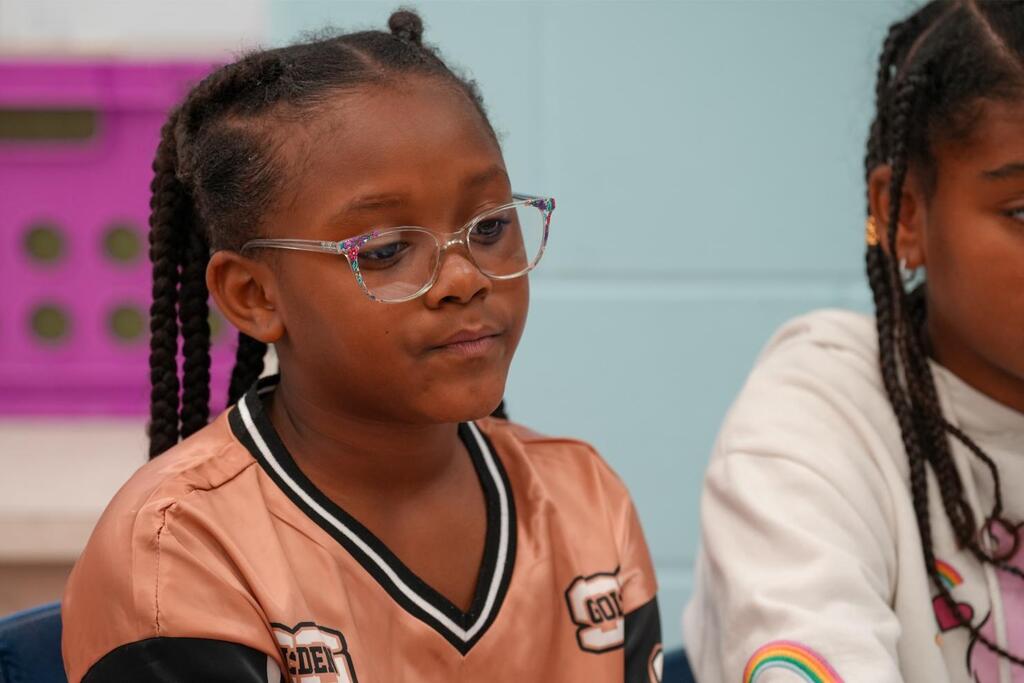 Two children sit in a classroom. The one wears glasses and a peach shirt with black trim. The other has braided hair and wears a white shirt.