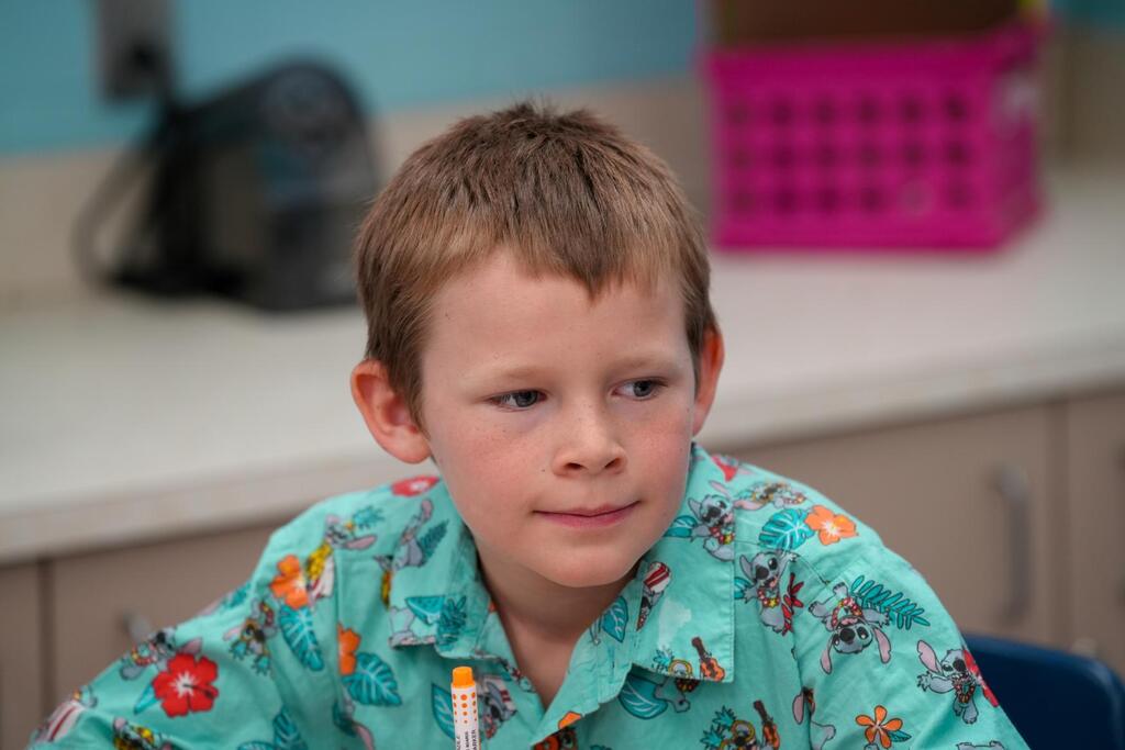 A young boy with short hair is seated at a table, wearing a green floral shirt. Behind him, there is a pink basket and a black bag.