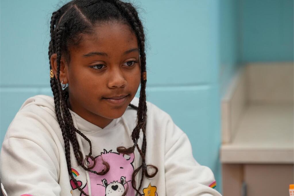A young girl with braided hair, wearing a white sweatshirt with a pink bear, sits in a room with blue walls.