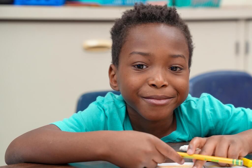 A young boy in a turquoise shirt sits at a table, smiling and holding a yellow pencil.