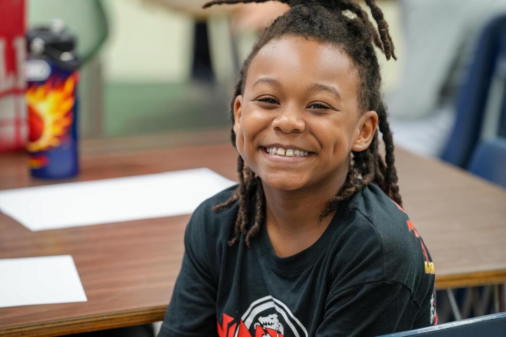A child with braided hair sits smiling at a desk, surrounded by a blue chair and table.