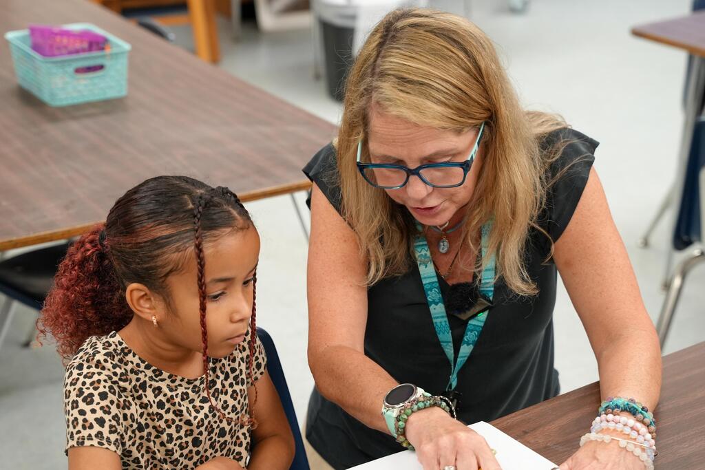 A woman wearing glasses and a black top helps a young girl with her work at a desk.