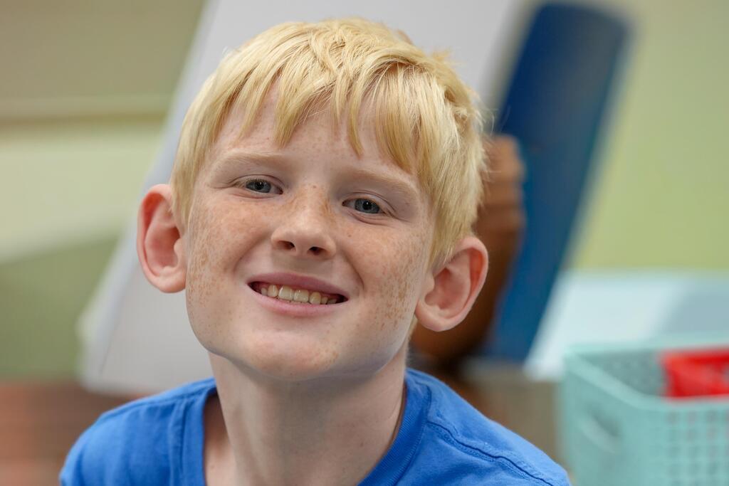 A smiling boy with blonde hair in a blue shirt. He is in a room with a green wall.