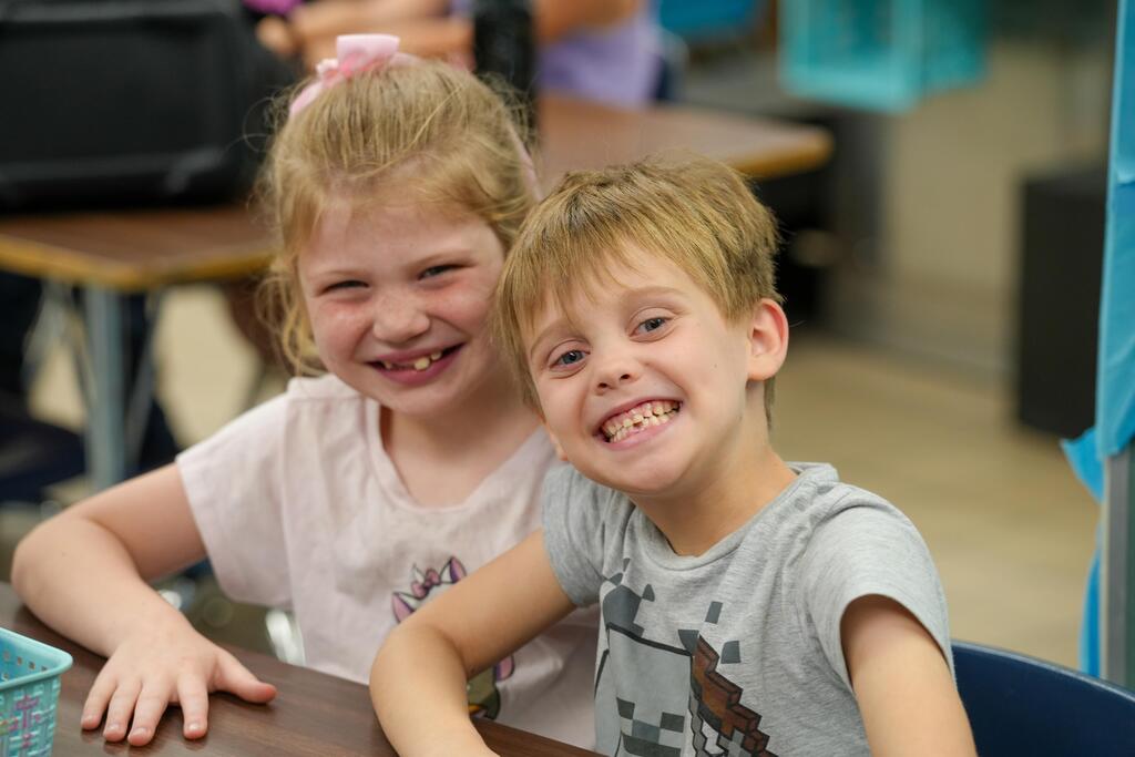 Two children sitting at a table, smiling. One is a girl with a pink top, the other a boy in gray.