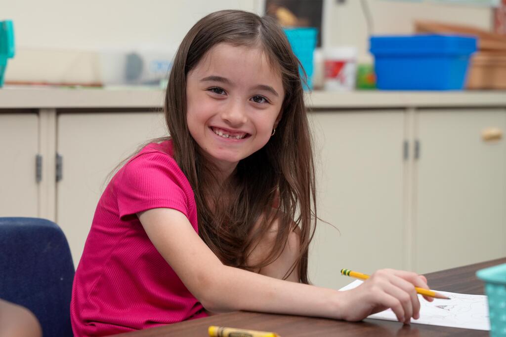 Girl with brown hair sits at a table holding a pencil. She smiles, with a pink shirt and a white paper.