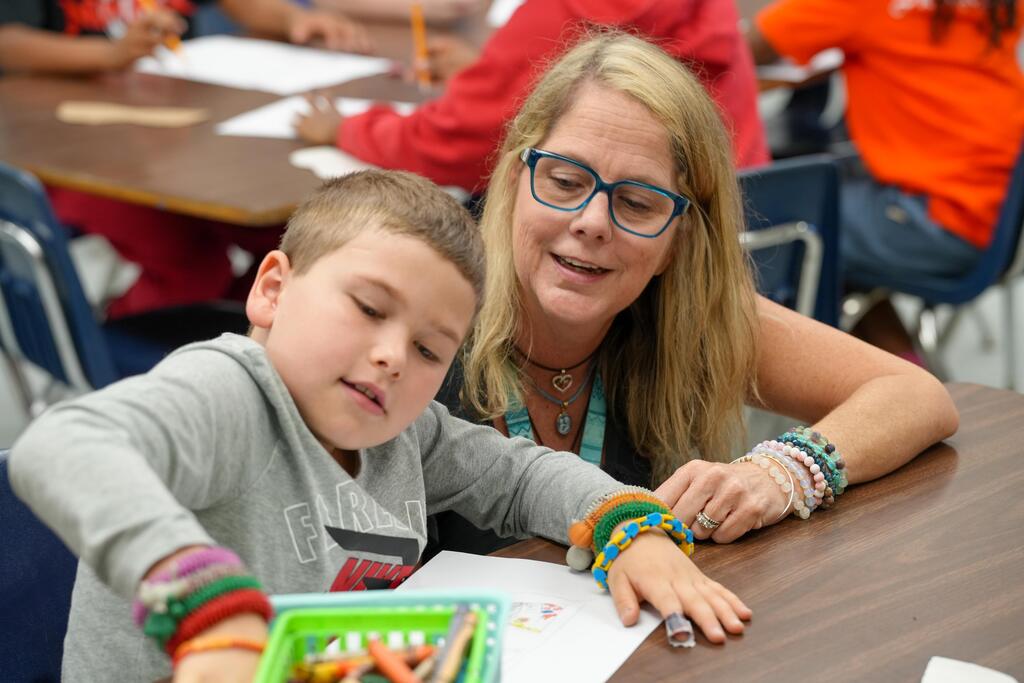 A woman with glasses and a necklace helps a child with bracelets and a pencil box at a table.