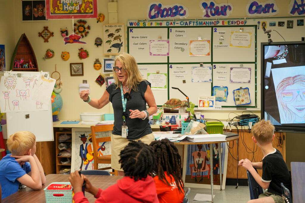 A teacher addresses a classroom of children; white board and a monitor visible in background.