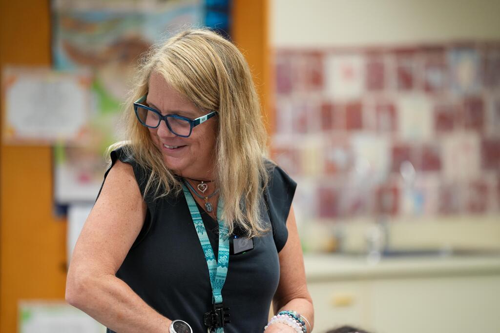 A woman with blonde hair and glasses stands indoors, wearing a black top and a blue lanyard. Background shows walls with educational posters.