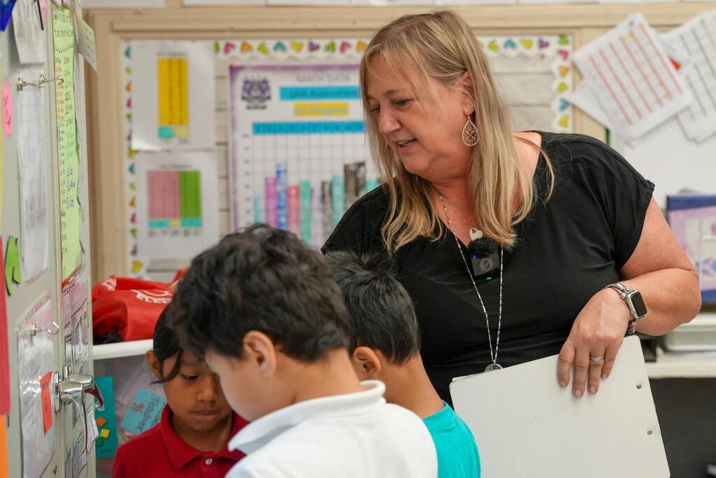 A woman stands behind two children in front of a classroom bulletin board. She holds a folder.