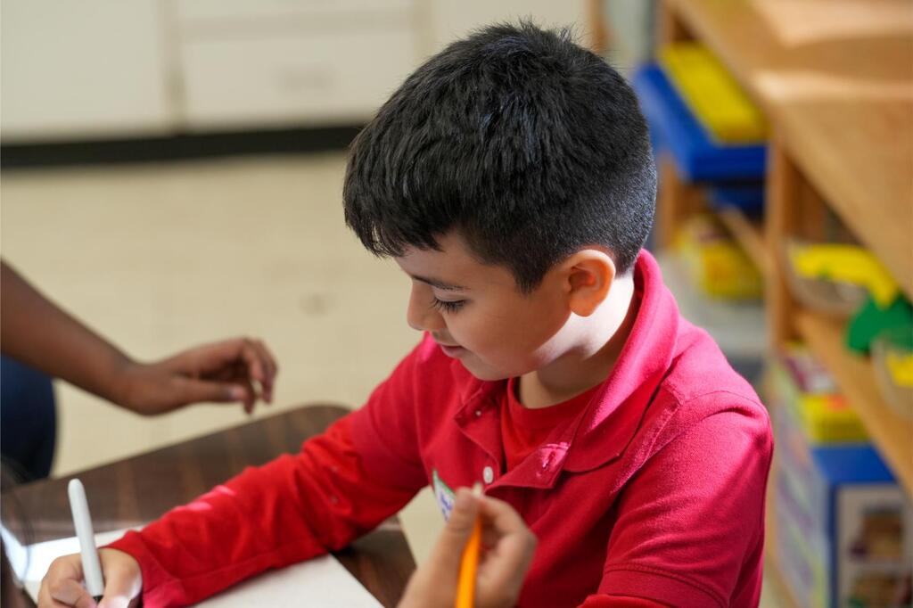 A young boy sits at a desk, writing on a paper with a pencil. He wears a red shirt.