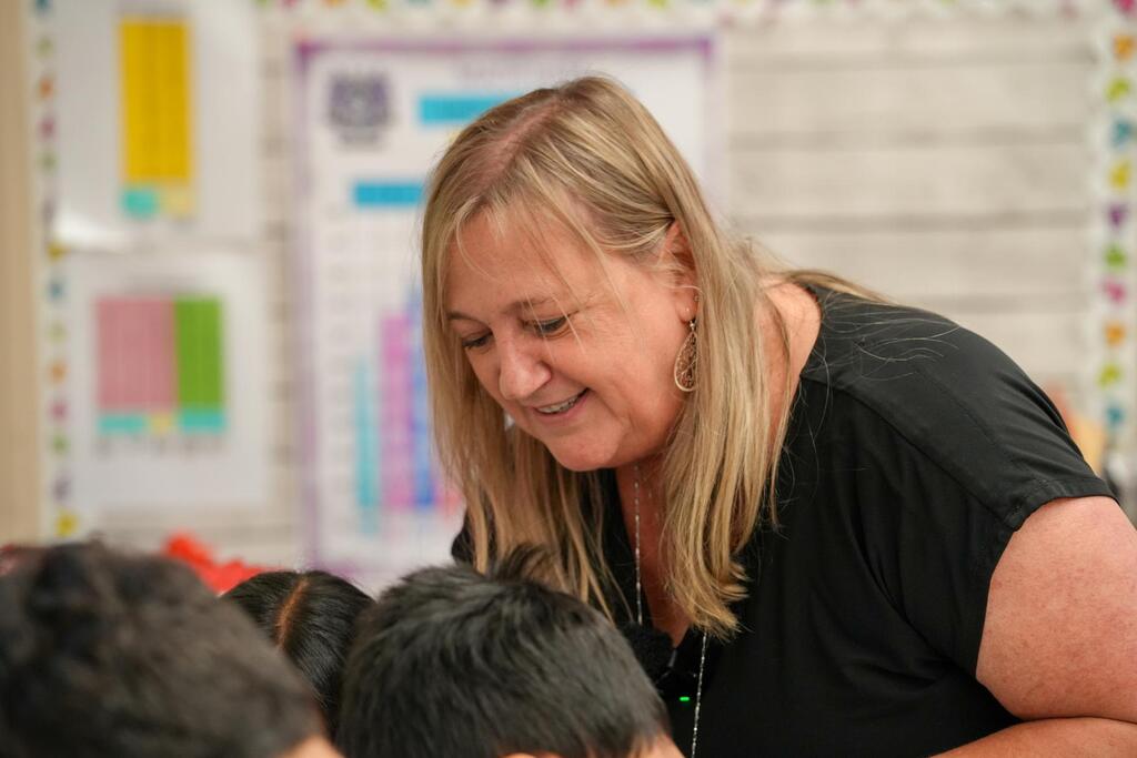 A woman with blonde hair bends over a group of children, all looking at her, in a classroom.