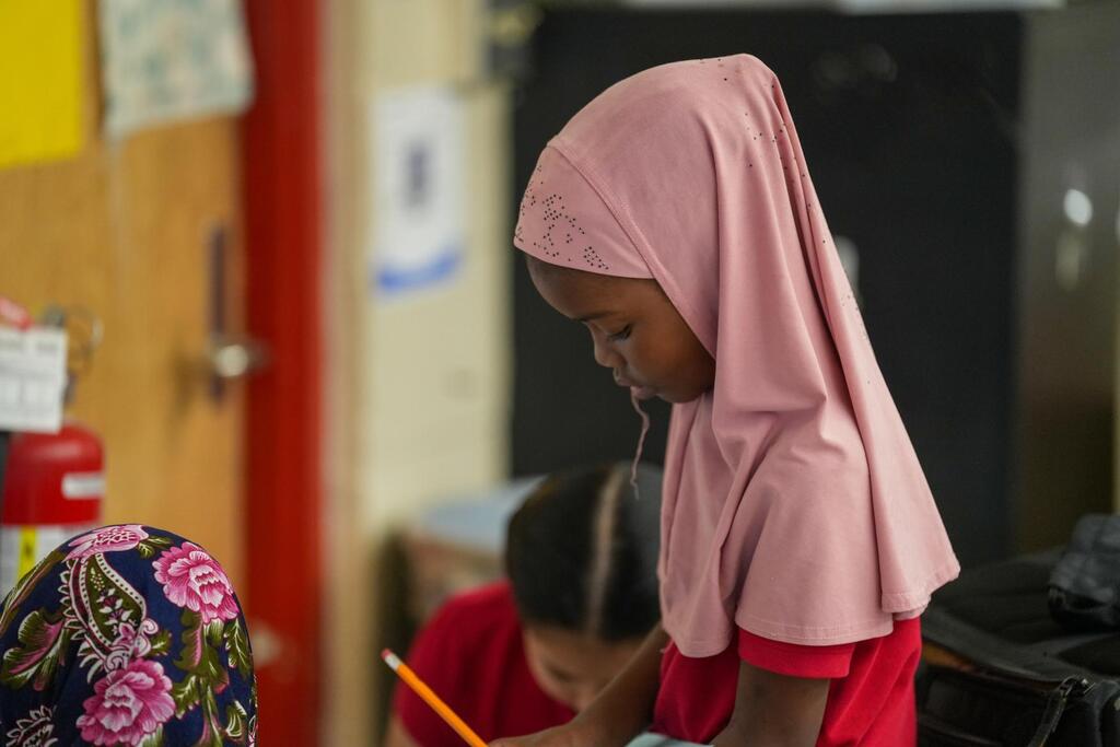 Girl in pink hijab and red shirt, writing in classroom. Another girl in floral head covering behind her.