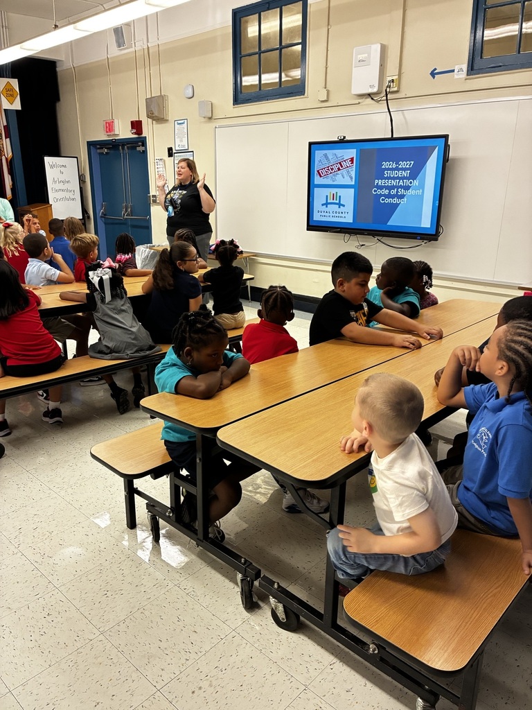 A teacher leads a lesson for young students seated at tables in a classroom. The room has a white tile floor, a television, and windows.