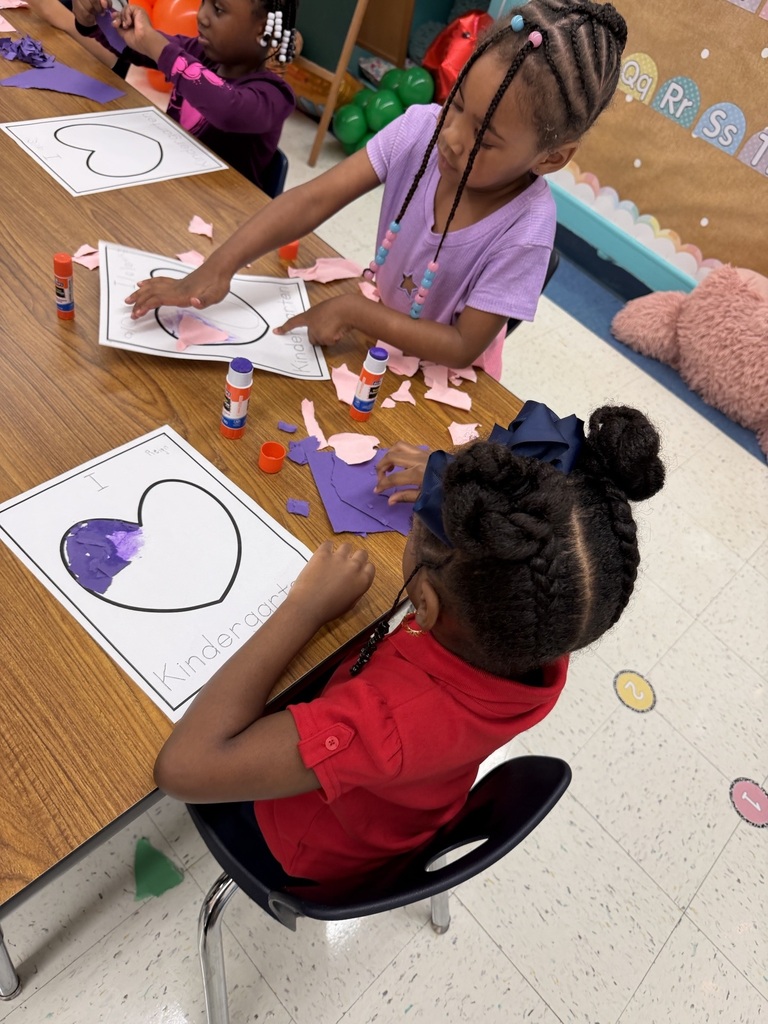 Children sitting at a table, crafting with paper and glue. One child works on a heart-shaped paper.