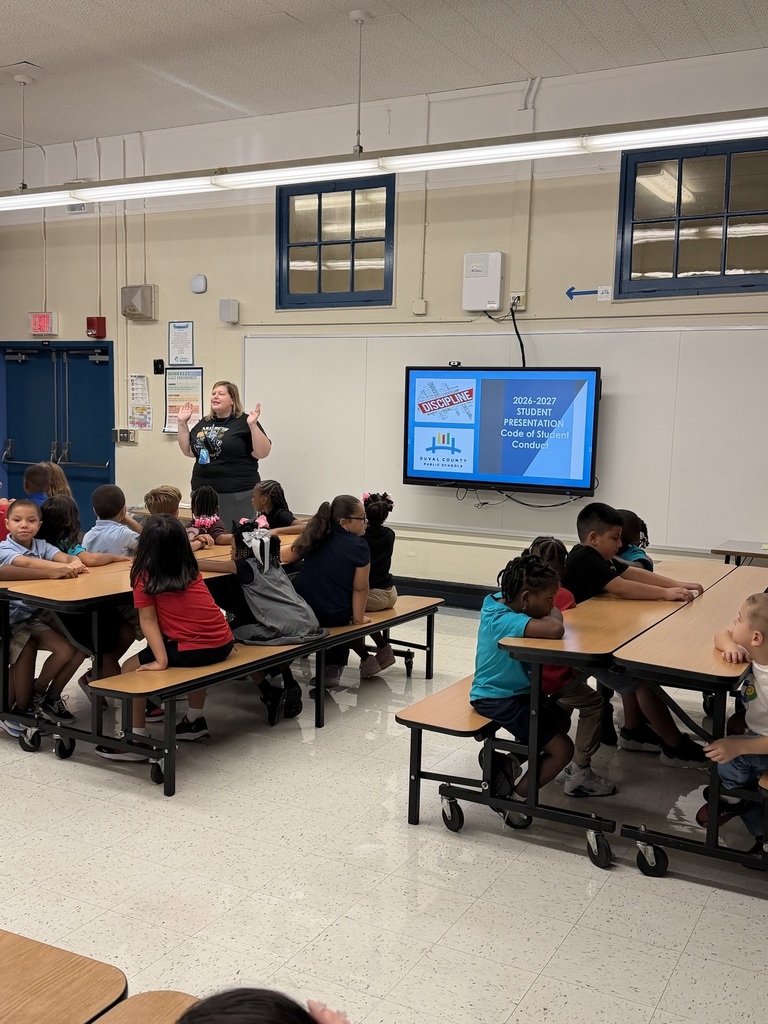 A classroom with students seated at desks. A teacher stands before a screen. White walls and windows.