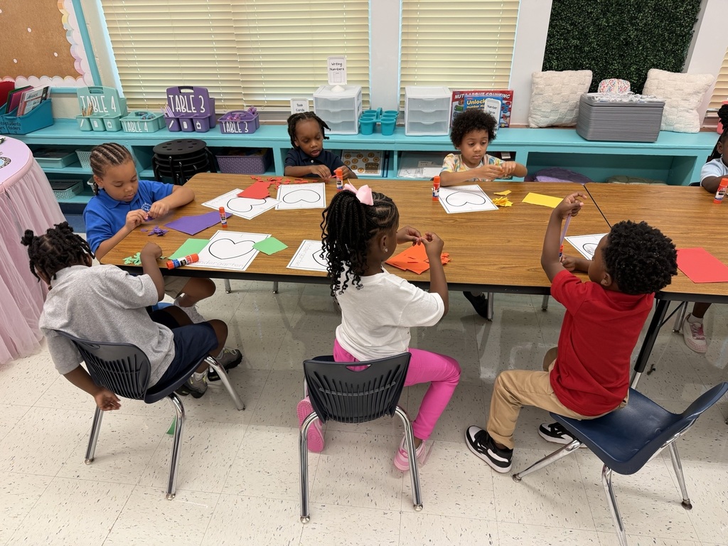 A group of children, wearing casual clothes, seated around a table in a classroom. They are engaged in a craft activity.