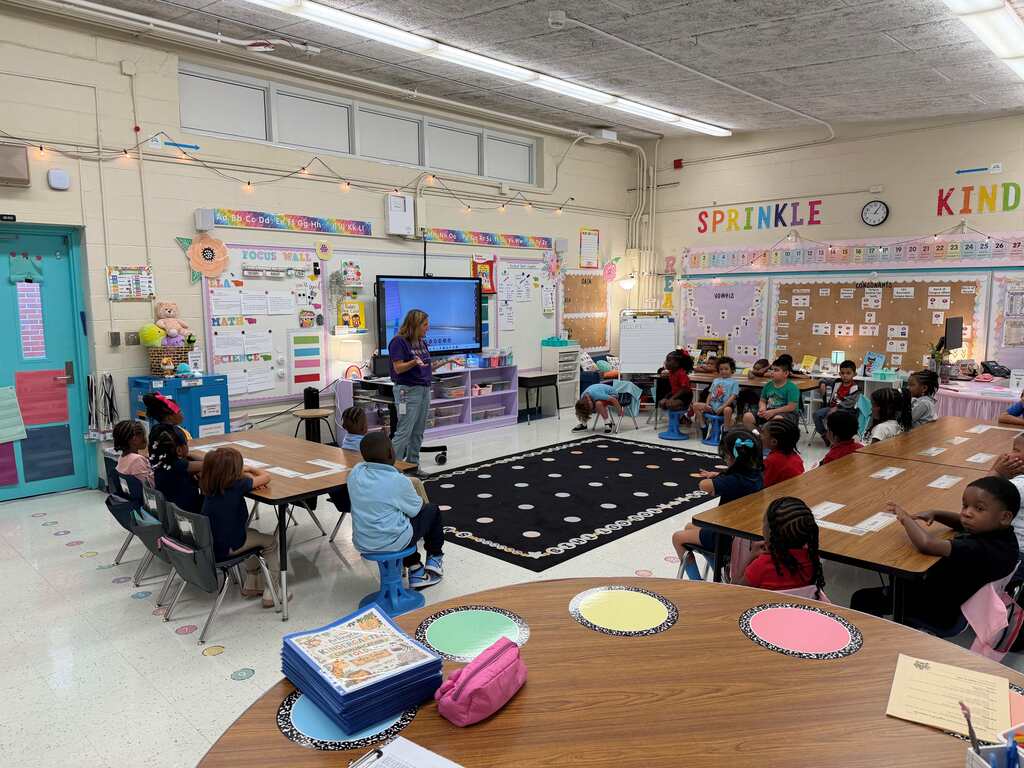 Classroom with a teacher standing in front of a screen, rows of students seated at desks, bulletin boards, and tables.