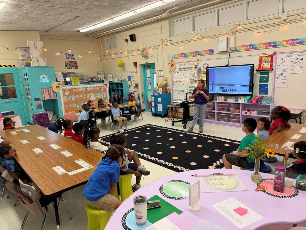 Kids sitting at tables in a classroom. A teacher stands before a class in front of a television.