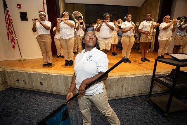 A student stands in front holding a flag, while a group of people in white shirts and khaki pants perform on a stage.
