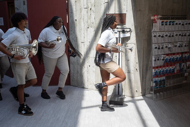 Three students in white shirts, shorts, and black shoes, playing trumpets. A red door and a white wall are in the background.