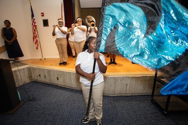 A student holds a large blue flag while standing on stage. Behind, a band plays trumpets. An American flag stands in the background.