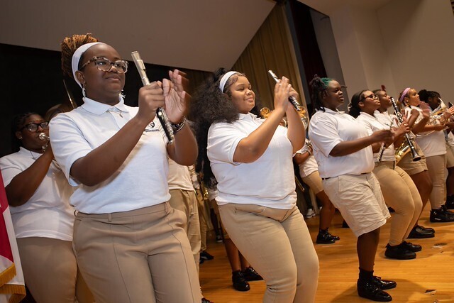 A group of students in matching white shirts and tan pants perform a coordinated routine with musical instruments.