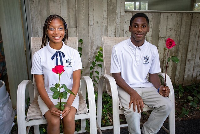 Two students in white polo shirts and light pants sitting on chairs. One holds a red rose. Behind them is a wooden wall and greenery.