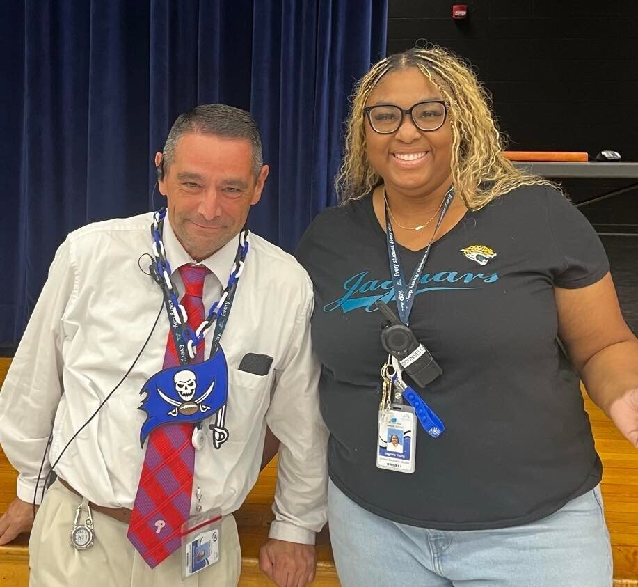 Dean Bombaro in a white shirt, red plaid tie and blue Buccaneer chain standing next to Counselor Jasmine Young in a black Jaguars t-shirt, smiling together on a school stage.