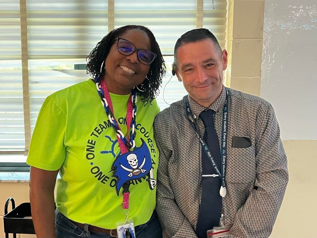 Two people in a classroom. Ms. Spruill wears a green shirt with a skull and crossbones. Dean Bombaro wears a gray shirt and a tie.