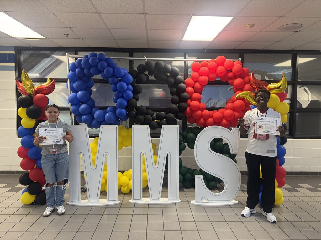 Two people pose in front of large letters and balloons. The letters spell "MMS".