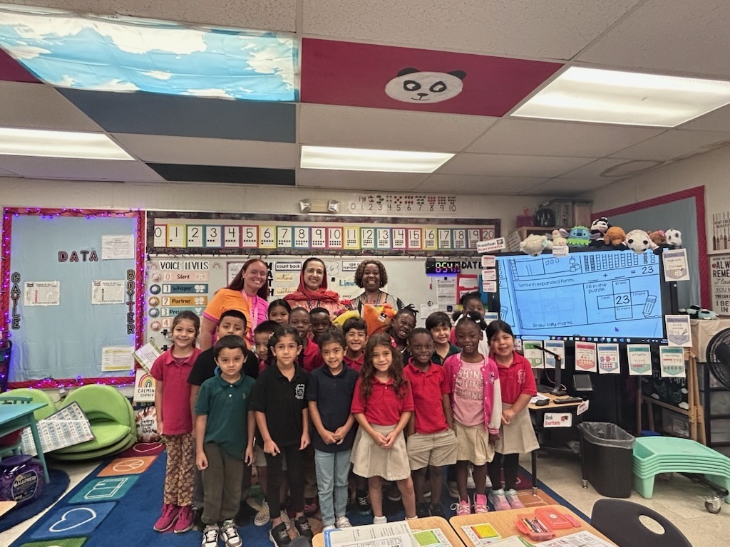 Classroom with a group of young children in red shirts. A teacher stands among them, a monitor on the wall, and a colorful ceiling.
