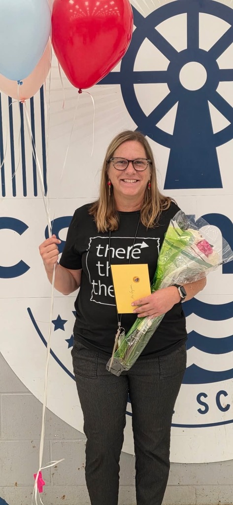 Woman in glasses and a black shirt holds balloons and a bouquet in front of a white sign.