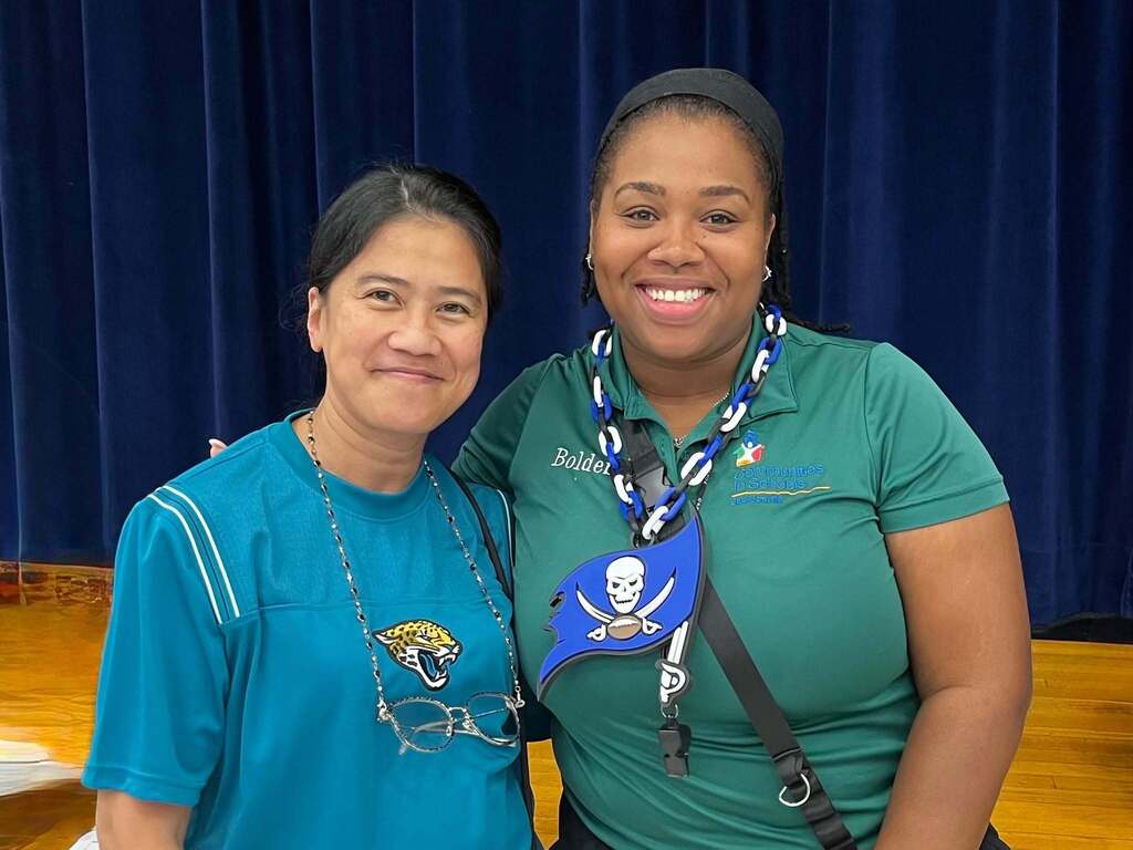 Two women, one in blue and the other in green, smiling, with a blue curtain in the background.